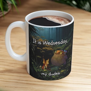 Peut inclure: Une tasse en céramique blanche avec une anse noire. La tasse présente un motif d'une créature verte et violette assise près d'un feu de camp dans une forêt sombre. Le texte "It is Wednesday, my dudes" est imprimé sur la tasse.