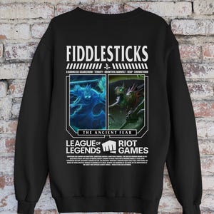 May include: Black sweatshirt featuring a graphic design with the text "FIDDLESTICKS" and "LEAGUE OF LEGENDS RIOT GAMES". The design includes two images and the text "THE ANCIENT FEAR". The sweatshirt also has the text "A HARMLESS SCARECROW - TERRIFY - BOUNTIFUL HARVEST - REAP - CROWSTORM".