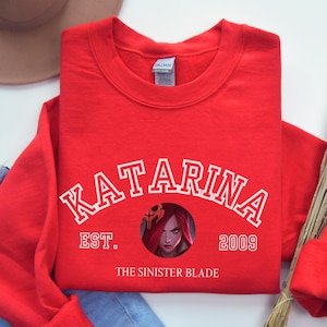 May include: A red crewneck sweatshirt with the word "KATARINA" in white arched lettering. Below the text is a circular graphic with a character illustration and the text "EST. 2009 THE SISTER BLADE". A brown hat and blue jeans are visible.