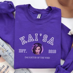 Puede incluir: Una sudadera morada con el nombre "KAI'SA" en letras blancas, un gráfico de un rostro femenino y el texto "EST. 2018 DAUGHTER OF THE VOID". La sudadera está doblada junto a unos vaqueros y una taza de café.