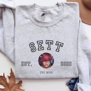 May include: A light gray sweatshirt with the word "SETT" in black block letters above a circular graphic of a person with pink hair. The text "EST. 2020" and "THE BOSS" are below the graphic. A brown hat and blue jeans are in the background.