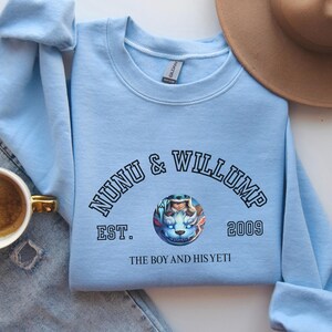 May include: A light blue sweatshirt with the text "NUNU & WILLUMP" arched above a circular graphic of a cartoon yeti. Below the graphic is "EST. 2009" and "THE BOY AND HIS YETI". A brown hat and a cup of coffee are also visible.
