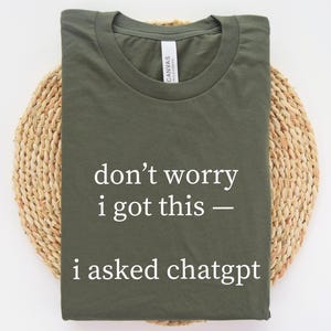 Könnte beinhalten: Olivgrünes T-Shirt mit dem weißen Text "don't worry i got this - i asked chatgpt". Das T-Shirt liegt gefaltet auf einem runden, gewebten, hellbraunen Platzset. Rundhalsausschnitt, kurze Ärmel.