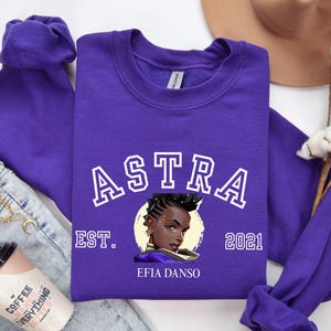May include: A vibrant purple sweatshirt featuring the word "ASTRA" in white, outlined letters. Below the word is a graphic of a woman's face. The text "EST. 2021" and "EFIA DANSO" are also printed on the sweatshirt.