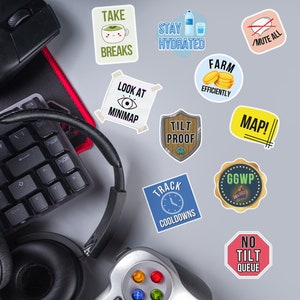 May include: A collection of gaming-themed stickers with phrases like "Take Breaks," "Stay Hydrated," and "Look at Minimap." Also includes a keyboard, mouse, headset, and game controller, all on a gray surface.