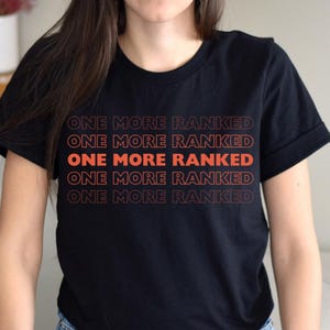 Può includere: T-shirt nera con la scritta arancione "ONE MORE RANKED" ripetuta cinque volte. Il testo è impilato verticalmente. La maglietta ha maniche corte e girocollo.
