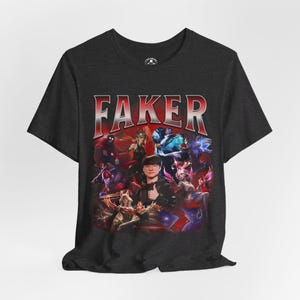 Op de afbeelding: Een donkergrijs T-shirt met een grafische print van een groep personages uit het videospel League of Legends. De tekst "FAKER" is in rode letters boven de grafische print gedrukt.