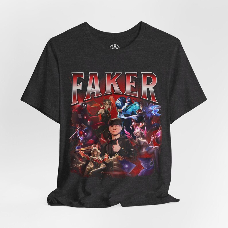 Op de afbeelding: Een donkergrijs T-shirt met een grafische print van een groep personages uit het videospel League of Legends. De tekst "FAKER" is in rode letters boven de grafische print gedrukt.