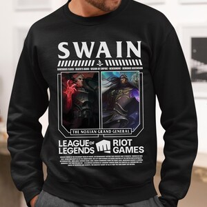 Puede incluir: Sudadera negra con el nombre "SWAIN" en blanco, sobre dos retratos de personajes. Debajo están las palabras "LEAGUE OF LEGENDS" y "RIOT GAMES". La sudadera tiene texto adicional.