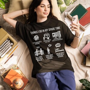 May include: Black t-shirt with the text "THINGS I DO IN MY SPARE TIME" and Dungeons & Dragons related icons. The shirt is worn by a person lying down, surrounded by books, a typewriter, and pillows. The person is holding a phone.