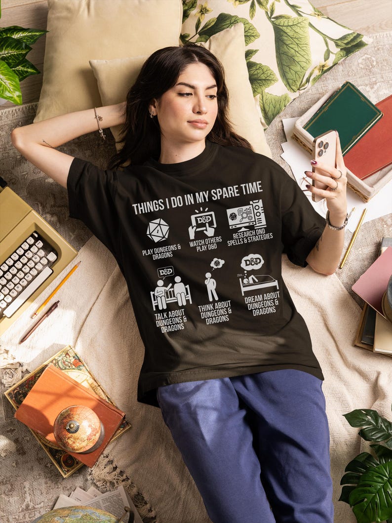 May include: Black t-shirt with the text "THINGS I DO IN MY SPARE TIME" and Dungeons & Dragons related icons. The shirt is worn by a person lying down, surrounded by books, a typewriter, and pillows. The person is holding a phone.