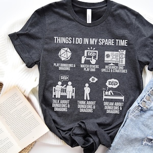 May include: Dark gray t-shirt with white text and graphics. The shirt reads "THINGS I DO IN MY SPARE TIME" with illustrations of dice, a screen, and people. The shirt's theme is Dungeons & Dragons.