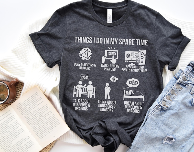 May include: Dark gray t-shirt with white text and graphics. The shirt reads "THINGS I DO IN MY SPARE TIME" with illustrations of dice, a screen, and people. The shirt's theme is Dungeons & Dragons.