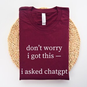 May include: A maroon t-shirt with the text "don't worry i got this - i asked chatgpt" in white. The shirt is laid on a woven, circular placemat.
