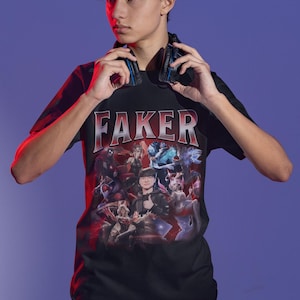Op de afbeelding: Een zwart T-shirt met een grafische print van een groep personages uit het videospel League of Legends. De tekst "FAKER" is in grote, vetgedrukte letters boven de grafische print gedrukt.