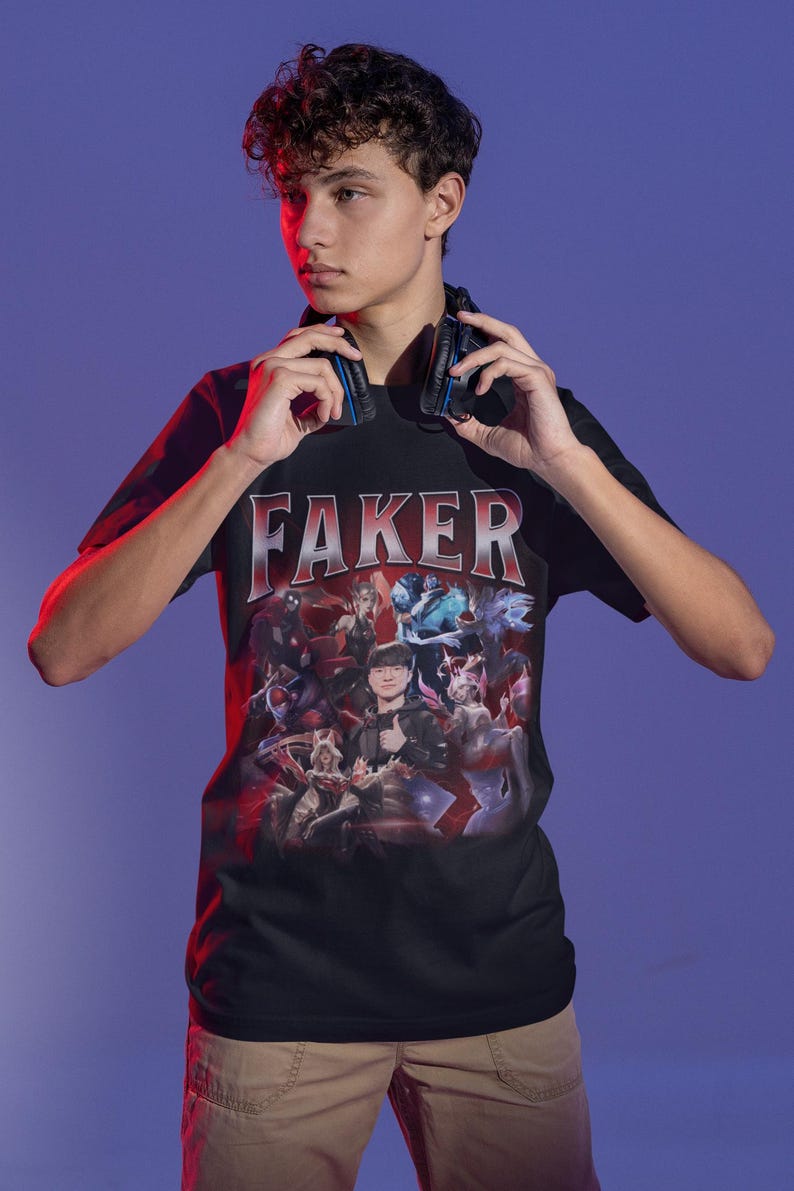 Op de afbeelding: Een zwart T-shirt met een grafische print van een groep personages uit het videospel League of Legends. De tekst "FAKER" is in grote, vetgedrukte letters boven de grafische print gedrukt.