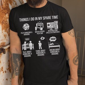 May include: Black t-shirt with white text and graphics. The shirt reads "THINGS I DO IN MY SPARE TIME" above six illustrations related to Dungeons & Dragons. The illustrations include a die, a D&D logo, and a person in bed.