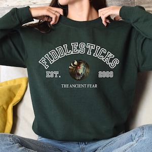 Puede incluir: Sudadera verde oscuro con el texto "FIDDLESTICKS" en letras blancas arqueadas sobre un gráfico circular. El gráfico presenta una cara de monstruo. Debajo del gráfico están las palabras "EST. 2009" y "THE ANCIENT FEAR" en blanco.