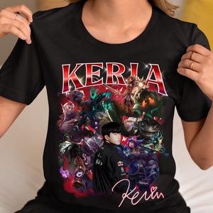 May include: Black t-shirt featuring a colorful graphic design with the word "KERLA" in red and white. The design includes various characters and the name "Kevin" in a cursive font. The shirt is a crew neck style.