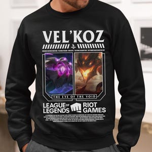 Könnte beinhalten: Schwarzes Sweatshirt mit dem Text "VEL'KOZ" und Bildern aus dem Videospiel League of Legends. Das Design enthält den Satz "THE EYE OF THE VOID" und die Logos von League of Legends und Riot Games.