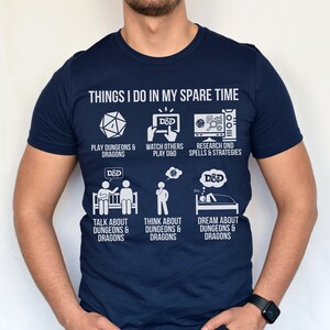 May include: Navy blue t-shirt with white text and graphics. The shirt reads "THINGS I DO IN MY SPARE TIME" with illustrations of activities like playing, watching, and dreaming about Dungeons & Dragons. A fun and thematic tee.