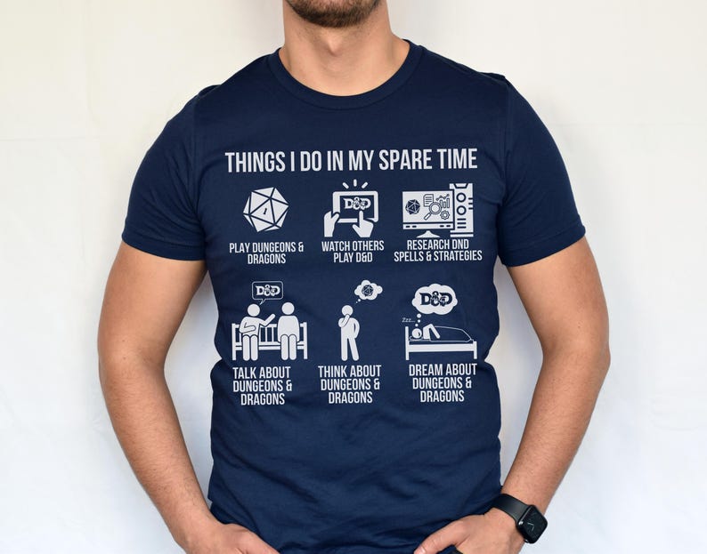 May include: Navy blue t-shirt with white text and graphics. The shirt reads "THINGS I DO IN MY SPARE TIME" with illustrations of activities like playing, watching, and dreaming about Dungeons & Dragons. A fun and thematic tee.