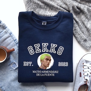 May include: A navy blue crew neck sweatshirt featuring the word "GEKKO" in white. Below the word is a circular graphic of a person with green hair. The text "EST. 2023" and the name "MATEO ARMENDÁRIZ DE LA FUENTE" are also printed on the sweatshirt.