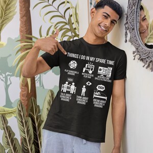 May include: Black t-shirt with the text "THINGS I DO IN MY SPARE TIME" and illustrations of activities related to Dungeons & Dragons. The shirt features icons and text describing various D&D-related pastimes.