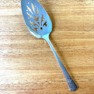 May include: Silver cake server with a decorative, openwork design on the blade. The handle has a floral pattern at the end. The server is on a wooden surface.