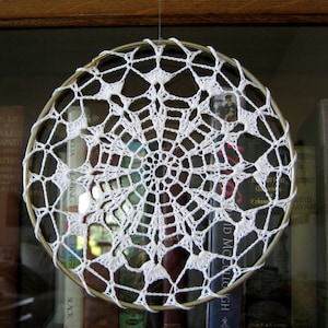 May include: A white crocheted dreamcatcher hanging from a wooden shelf. The dreamcatcher is circular and has a delicate, intricate design.