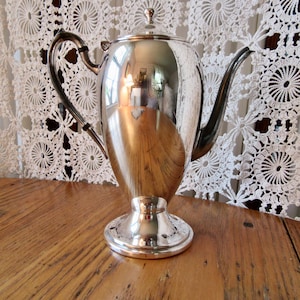 Könnte beinhalten: Eine silberne Teekanne mit einem runden Korpus und einem hohen, schlanken Ausguss. Die Teekanne hat eine polierte Oberfläche und einen dekorativen Griff.