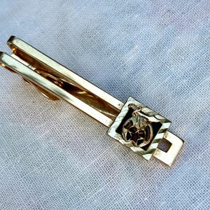 May include: A gold-toned tie clip with a decorative square element. The square features a black background with a gold emblem and a red star. The clip is designed to secure a tie.