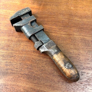 May include: An antique adjustable wrench with a dark, weathered metal head and a wooden handle. The wrench features a screw mechanism for adjusting the jaw size. The handle is a light brown color with a circular metal accent.