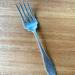 May include: A silver fork with four tines, resting on a wooden surface. The handle features an embossed floral design. The fork is a vintage piece, likely for dining.