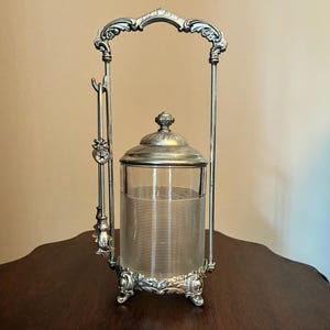 Op de afbeelding: Een verzilverde kruidenhouder met een heldere glazen pot met horizontale ribbels. De pot heeft een bijpassend zilveren deksel en staat in een decoratief metalen frame met een boogvormig handvat en een kleine bijbehorende zilveren lepel.