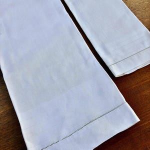 May include: Two white linen towels with a textured weave. The towels have a decorative border with a stitched edge. One towel is folded, and the other is unfolded, showing the full length.