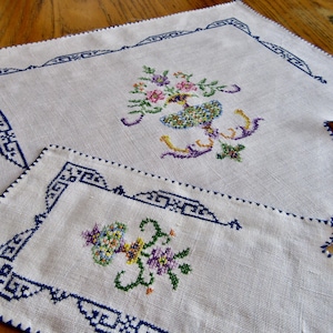 May include: Two white linen placemats with blue embroidered borders and floral designs. The larger placemat has a vase with flowers in the center, while the smaller placemat has a similar design with a smaller vase.