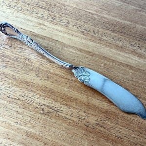 Puede incluir: Cuchillo de mantequilla plateado con detalles florales ornamentados en el mango y la base de la hoja. La hoja es curva y ancha, con una punta redondeada. El mango presenta un diseño decorativo. El cuchillo está sobre una superficie de madera.