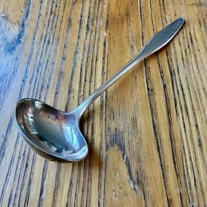 May include: A silver-toned metal ladle featuring a deep, rounded bowl and a long, slender handle that tapers to a point. The surface of the ladle is smooth and reflective, showing a polished metallic finish throughout the entire utensil.