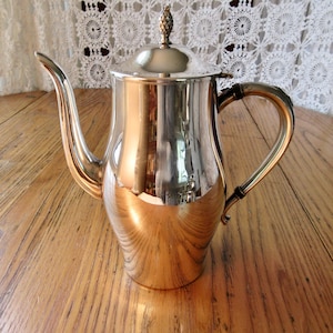 Puede incluir: Una tetera de plata con un acabado brillante y un pomo decorativo en la tapa. La tetera tiene un cuerpo curvo y un pico largo.