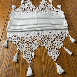 May include: White lace table runner with a scalloped edge and decorative tassels. The runner is made of a folded fabric with an intricate lace design along the top and bottom edges. The tassels are attached at intervals along the edges.