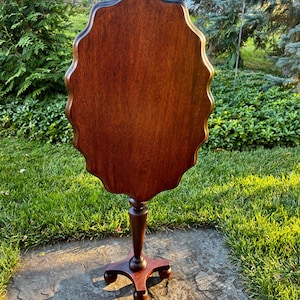 May include: A dark brown wooden pedestal table featuring a scalloped, oval-shaped tabletop surface. The table is supported by a single central column that transitions into a four-legged base, with each leg ending in a rounded, ball-shaped foot.