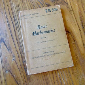 Puede incluir: Un libro de color beige titulado "Basic Mathematics" con el texto "Education Manual EM 308" en la parte superior y "Reprinted for The United States Armed Forces Institute" en la parte inferior.