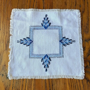 Op de afbeelding: Een vierkante witte stoffen onderzetter of kleine mat met een centrale vierkante rand geborduurd in licht- en donkerblauw garen. Vier symmetrische blauwe bloemachtige motieven strekken zich uit vanuit het midden en de randen zijn afgewerkt.