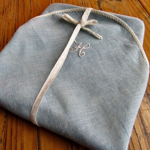 May include: A folded, pale blue handkerchief with a cream ribbon tied around it. The handkerchief has a cream trim and an embroidered "H" initial. The item is presented on a wooden surface.