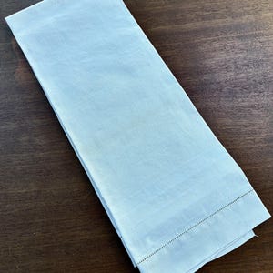 Può includere: Un tovagliolo di lino blu chiaro piegato, con una trama strutturata. Il tovagliolo ha un bordo orlato con una linea di piccole cuciture scure. Il tovagliolo è rettangolare e si trova su una superficie di legno marrone scuro.