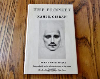 May include: A hardcover book titled "THE PROPHET" by Kahlil Gibran. The cover features a portrait and the text "GIBRAN'S MASTERPIECE". The book is published by Alfred A. Knopf, New York. The book is on a wooden surface.