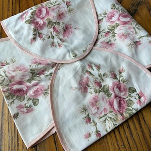 Puede incluir: Cuatro servilletas de tela blancas con un estampado floral de rosas en tonos rosa y verde. Cada servilleta tiene un ribete rosa y un borde curvo. Las servilletas están dispuestas sobre una superficie de madera.