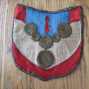May include: A shield-shaped embroidered patch with a red border and accents. The top section is blue with a red teardrop shape. The center features spiraling, circular embellishments in a darker color. The background is a tan color.