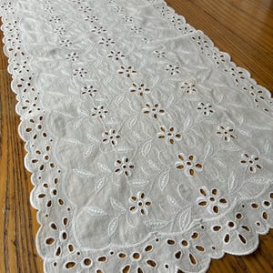 May include: White eyelet table runner with a scalloped edge. The runner features embroidered floral and leaf patterns throughout. The delicate design adds a touch of elegance to any table setting. The runner is made of a lightweight fabric.